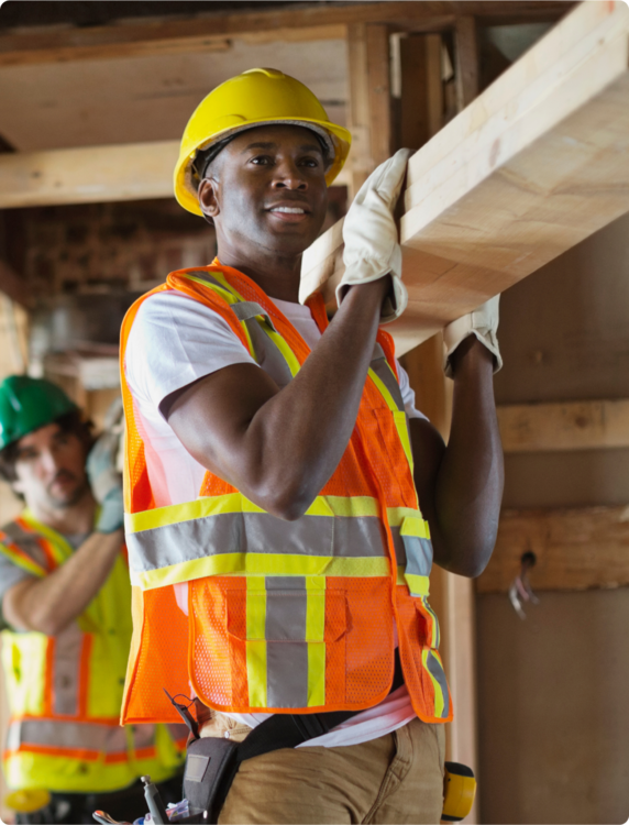 Two men wearing yellow hard hats and construction safety vests carrying a long piece of lumber.