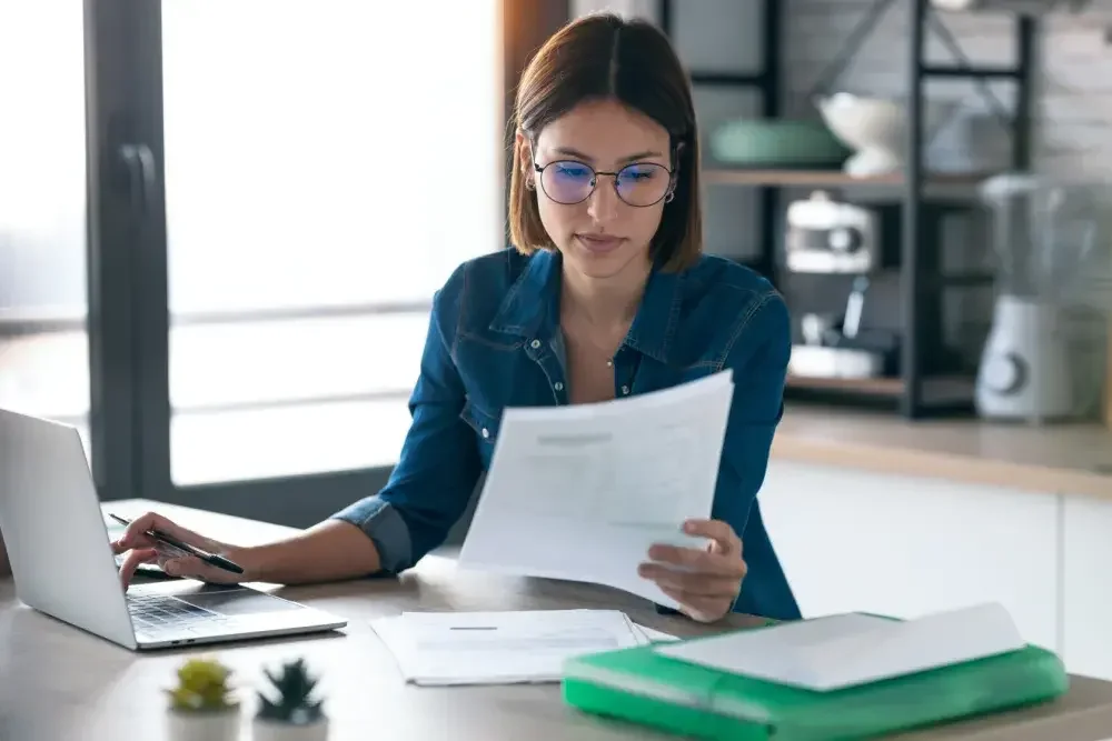 A woman seated at a desk holds documents in one hand while creating a logo for her new business on her laptop with the other hand. Some new businesses will have to get a federal tax ID number (FEIN).