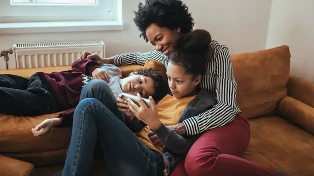 A woman smiles and lounges on the couch with her children while they all watch a video on a phone.