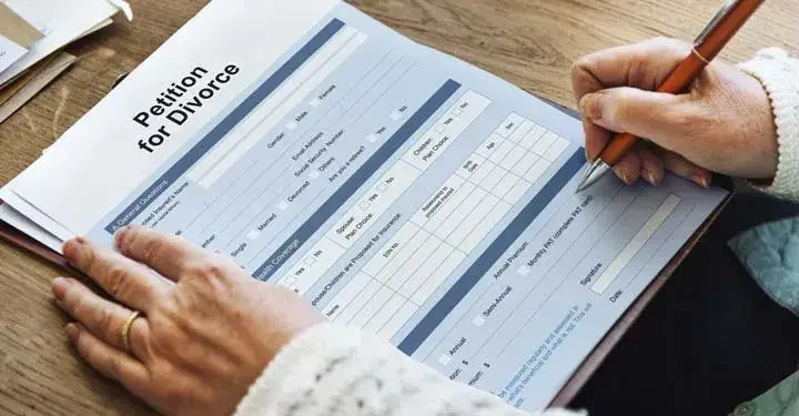 Wrinkled hands in white fluffy sweater with gold wedding ring filling out "petition for divorce" with ballpoint pen