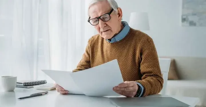 Old man in black glasses, blue button down, black watch, and yellow sweater looking at two sheets of paper on white desk