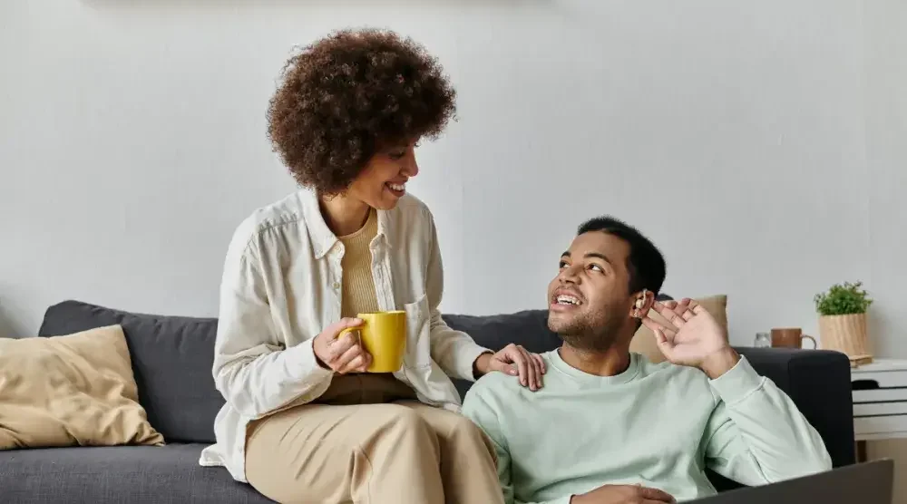 A woman sitting on a couch and holding a coffee cup smiles and puts her hand on the shoulder of her common-law husband, who motions to his hearing aid and is seated on the floor.