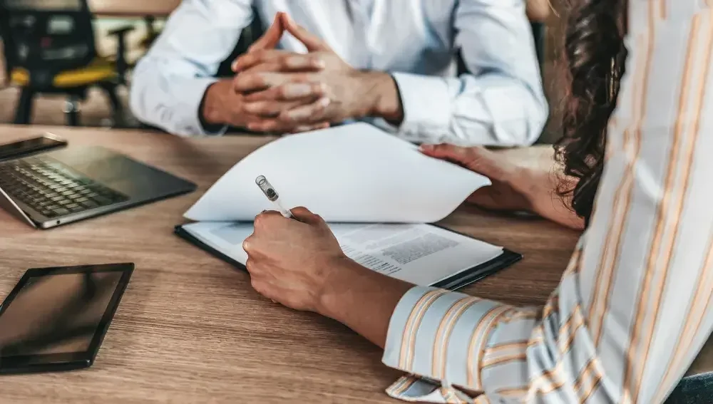 A person signs papers while someone else looks on. A release of liability form is designed to safeguard you against lawsuits by planning for and addressing potential disputes.