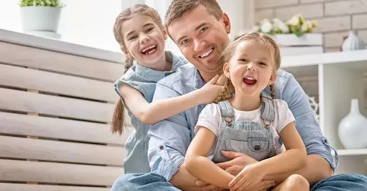 Smiling dad sitting with his two laughing children in matching light wash denim in living room