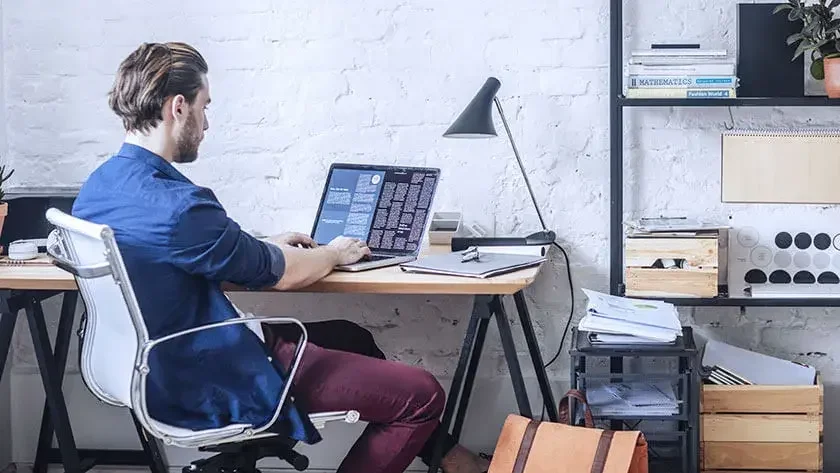 man-sitting-in-home-office-using-laptop wearing blue shirt 