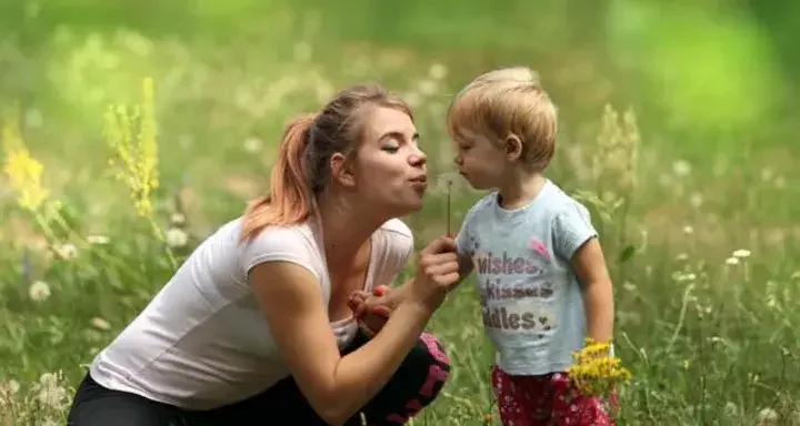 Woman and toddler sit in green field and blow on a dandelion together