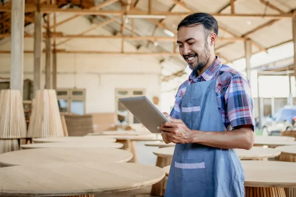 A man looks at an email from his registered agent on a tablet in his furniture store, where he is surrounded by wooden tables and other items. Every LLC in the state of Florida is required to name a registered agent.