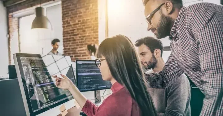 Three people gathering in office around two desktop computers displaying code