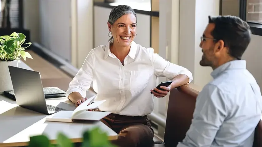 A smiling attorney holds a smart phone and sits at a conference table in an office with a new business owner as he consults her about forming a business entity. 