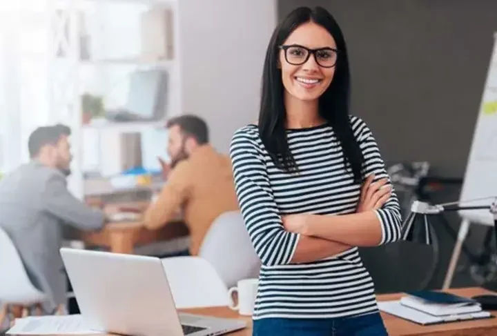 Woman crossing arms and smiling in office in front of two businessmen talking and desk with laptop and coffee cup on it