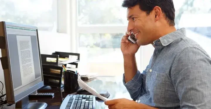 Man smiling while on landline phone holding papers and looking at document on desktop computer