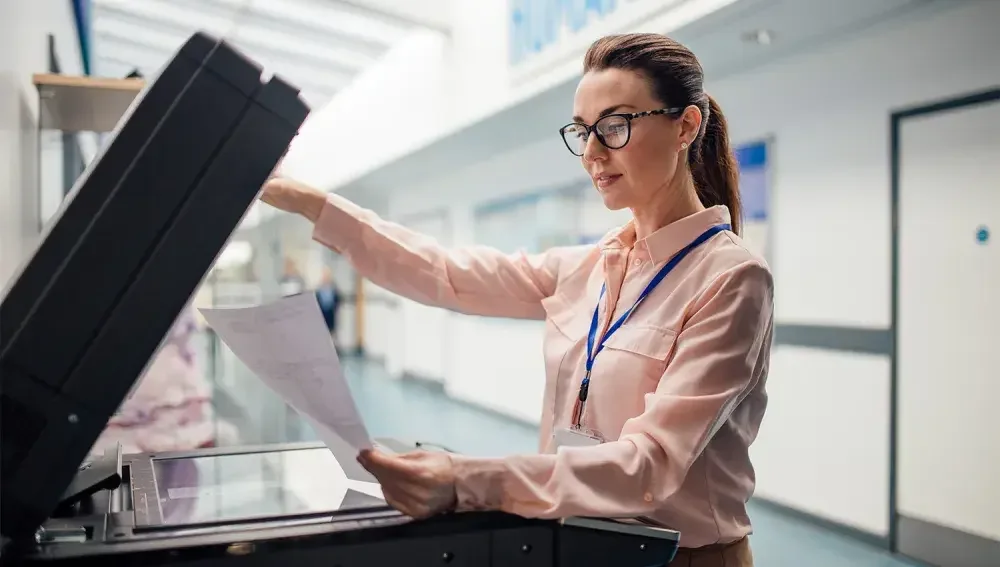 A woman with brown hair, glasses, a light pink shirt and a blue lanyard around her neck puts a document into a large scanner.