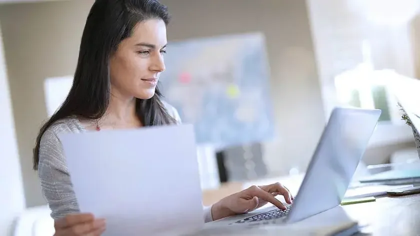 A woman sits at a desk with a piece of paper in one hand and types on a laptop computer with the other hand.