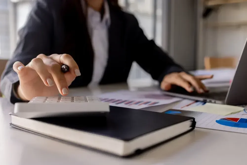 A woman seated at a desk uses a calculator with one hand while typing on her laptop with her other hand.