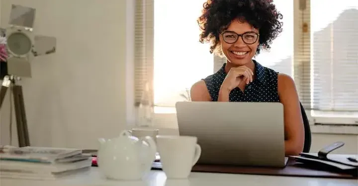 Woman in home office pauses from work to smile with hand under chin with tea kettle in front of her laptop