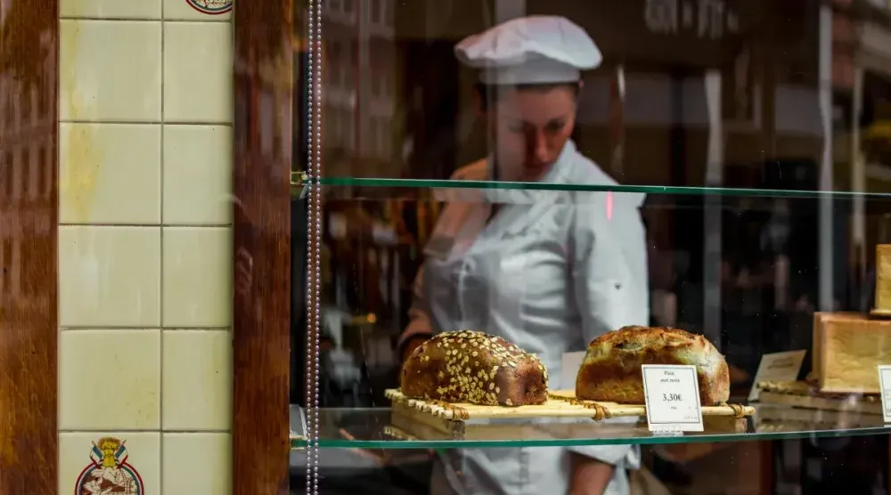 A female baker in uniform inspects a shelf of bread loaves displayed in a bakery storefront window.