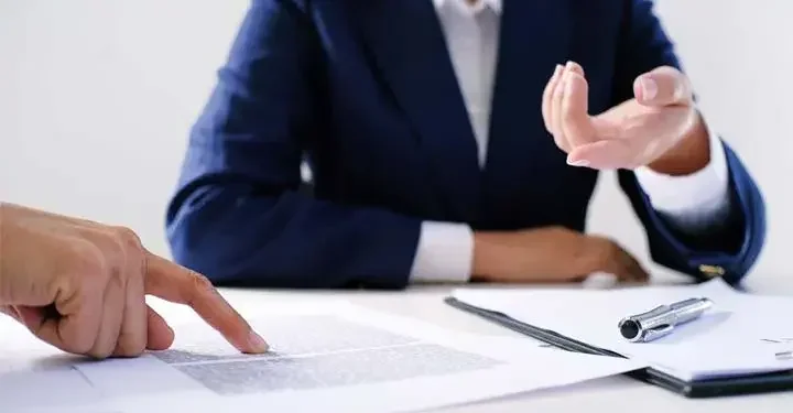 Hand points to text on sheet of paper resting next to stack of paper with pen on it as other person gestures towards papers