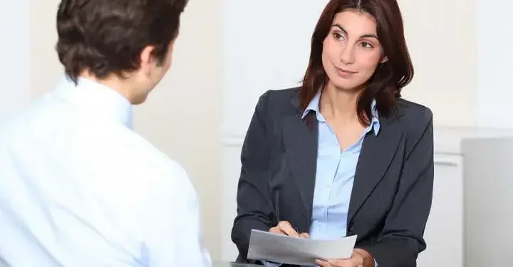 Woman raising eyebrows while writing on sheet of paper and looking at man facing her