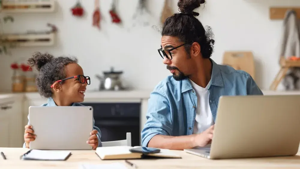 A child and father, both wearing eyeglasses, sit at a counter as the child holds an iPad and the father works on a laptop computer. It's important to have a will if you have children under the age of 18.