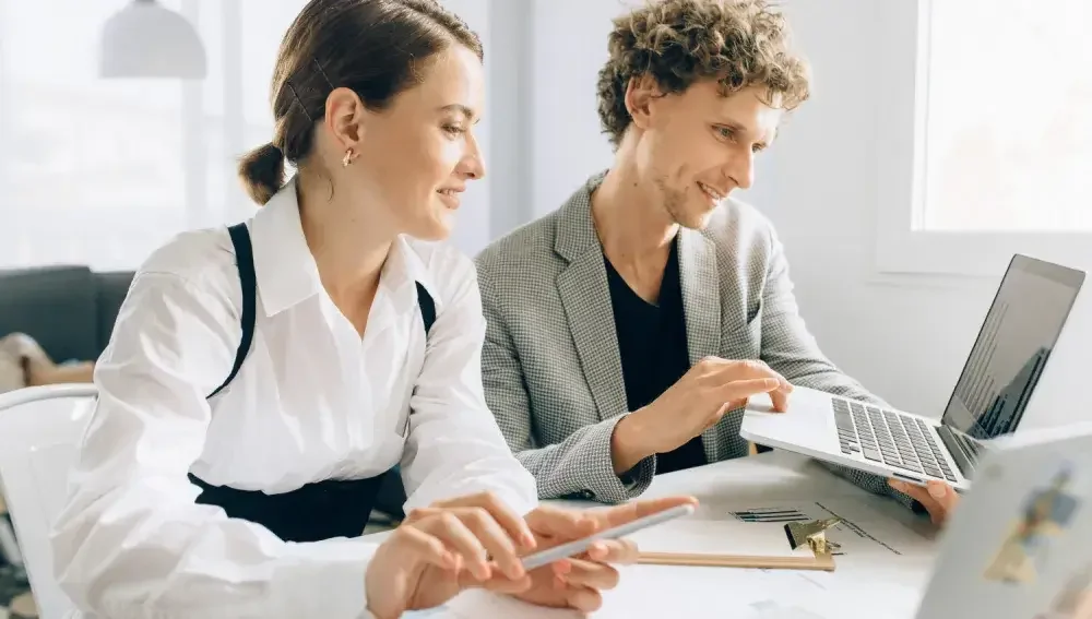 A woman and man seated at a table look at the screen of an open laptop. Learn the steps to set yourself up as a consulting firm.