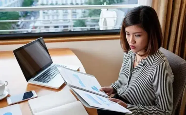 Woman at office desk with laptop, phone, and coffee cup resting on it and reading graphs on papers in a folder while sitting next to a window showing busy streets below