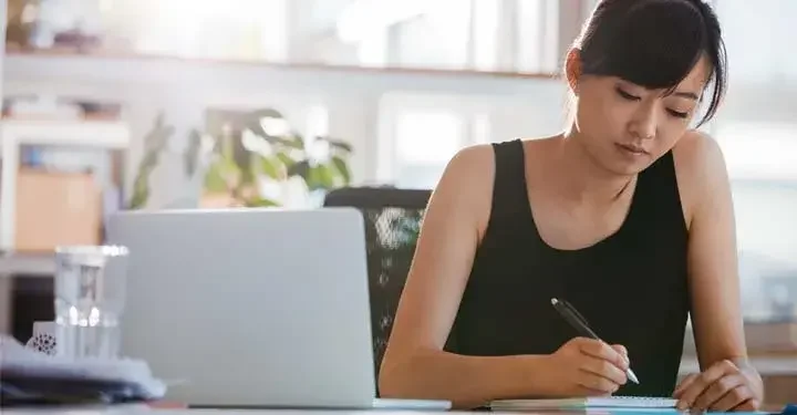 Woman in office in black tank top writing in notebook in front of laptop