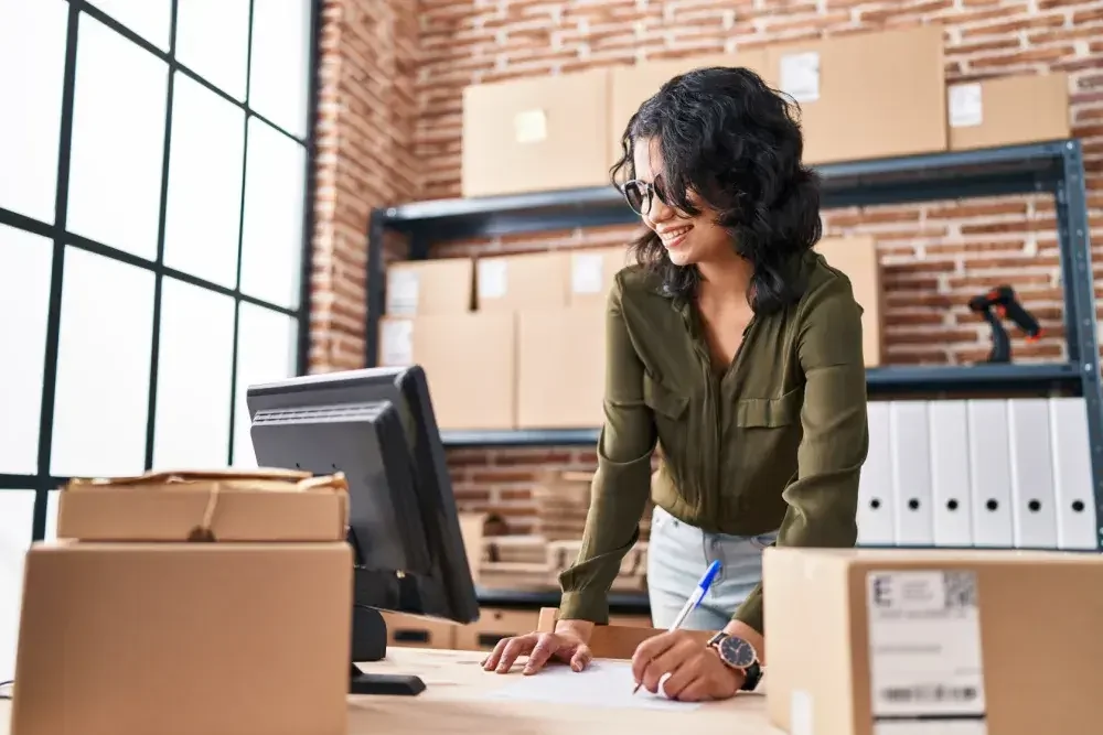 A woman stands at her desk taking notes in front of her computer as she opens an LLC bank account. Learn how to open LLC bank account.