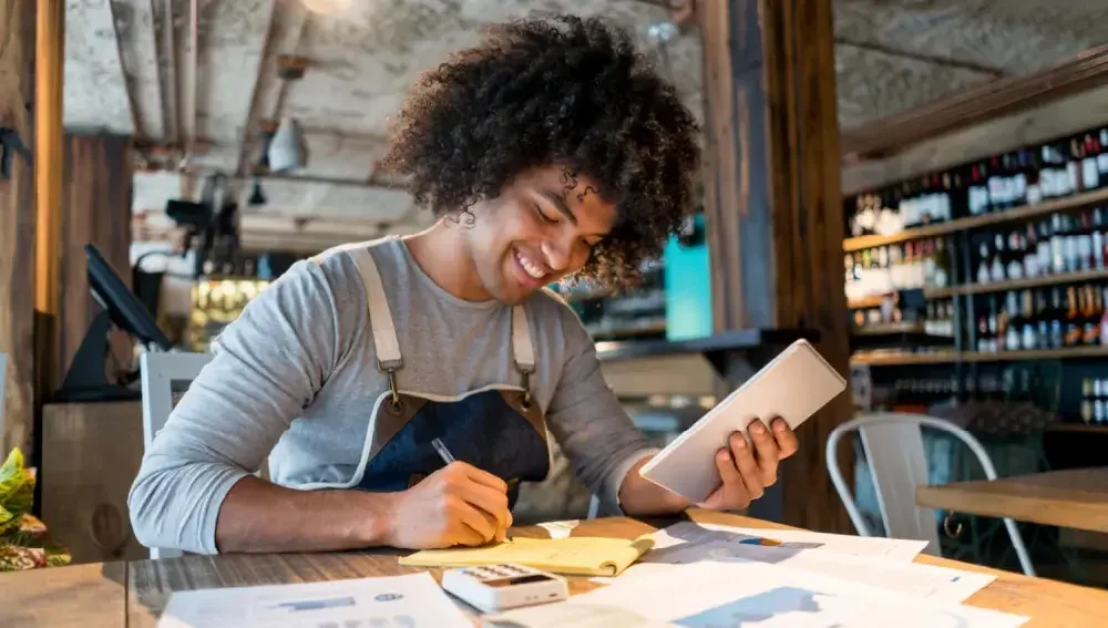 A man in an apron seated at a table takes notes with one hand while looking at an iPad or other smart electronic device with the other hand. Learn foolproof tips to help you easily manage business expenses..