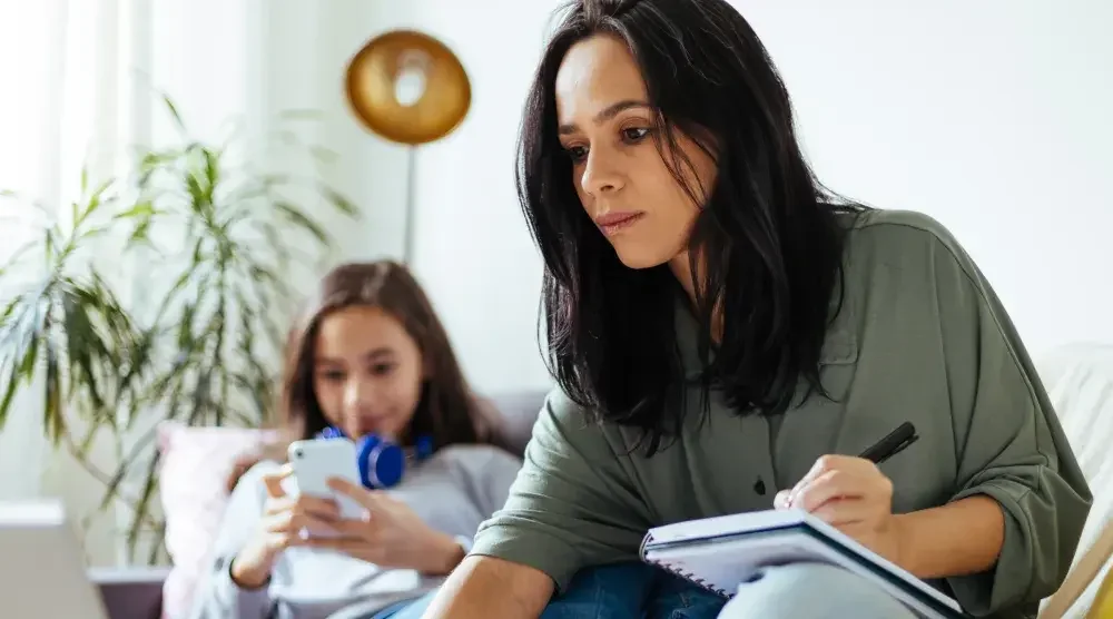 A soon-to-be-divorced mother takes notes while looking at her laptop computer. Behind her, a child wearing headphones around her neck looks at a cell phone. Spousal support can be one of the most complicated and contentious parts of a separation or divorce. An experienced spousal support lawyer can guide you through the process.