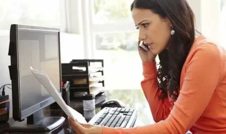 Concerned woman talking on the phone while looking at paper at her desk in front of her desktop computer