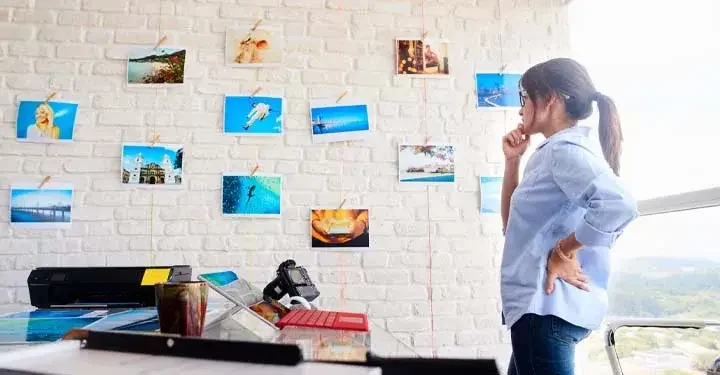 Woman in blue button up shirt with hand on hip looking at photos on white brick wall in office