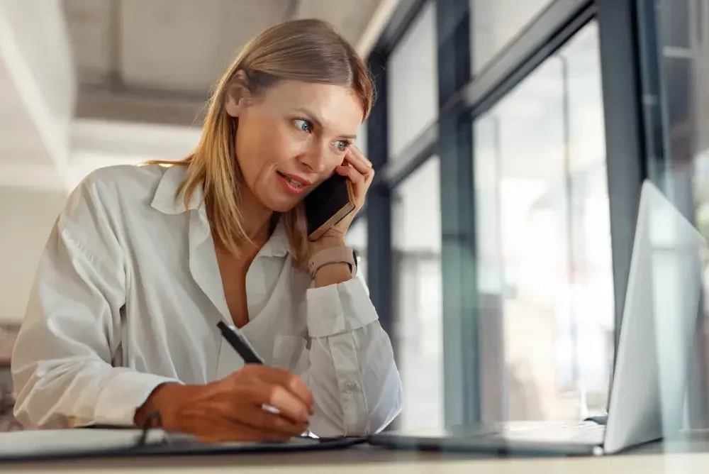 A woman seated at a desk in an office talks on her cellphone to a trademark attorney. The company's trademark is a legal protection given to a company or brand. 