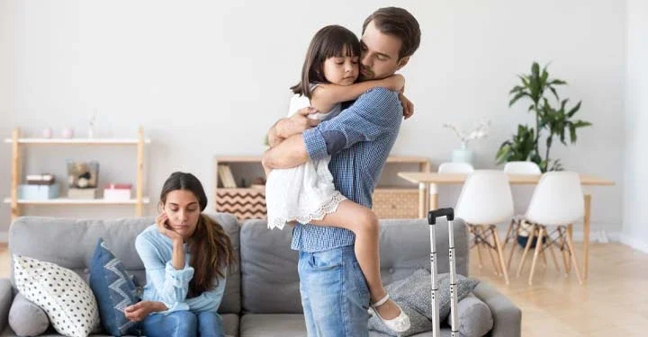 Mother sitting solemnly on a couch while a father and daughter hug next to a suitcase