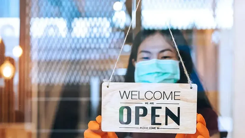 woman-wearing-mask-holding-open-sign-in-store-window