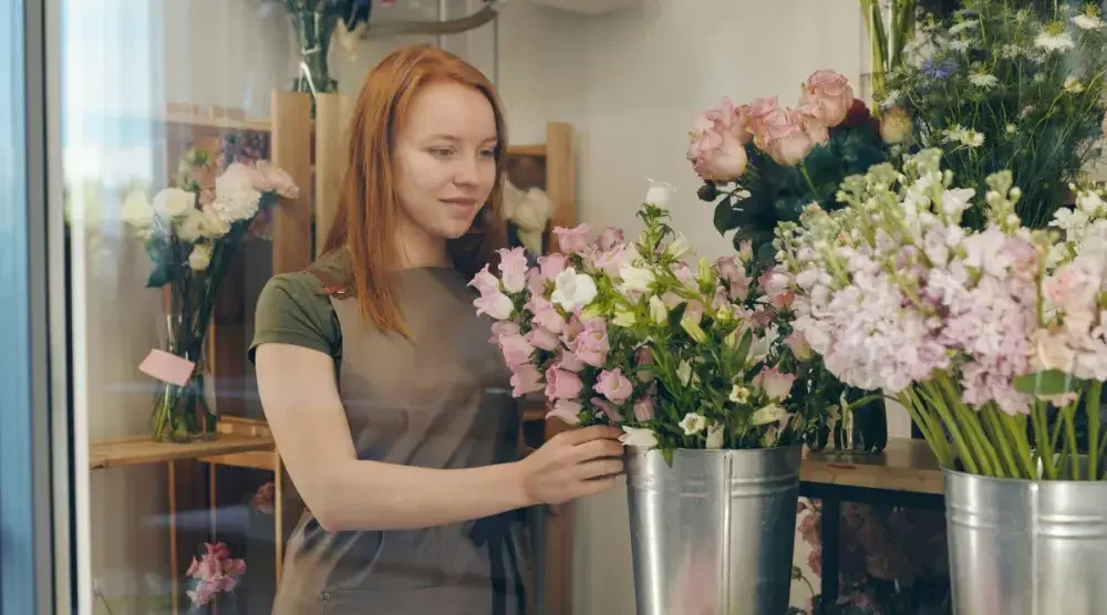 A female redheaded florist looks at an arrangement of pink and white roses in her flower shop with a satisfied smile.