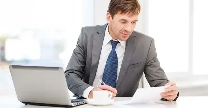 Man holding a piece of paper and pen working at a desk with a laptop and cup of coffee