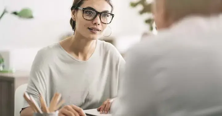 Woman in grey sweater and chunky glasses listens as out of focus businessperson talks