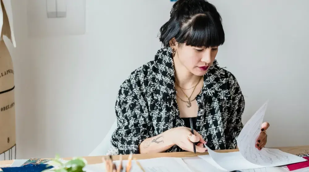 An artist wearing a large plaid scarf sits at a desk in her bright, airy studio and reviews paperwork for her LLC.