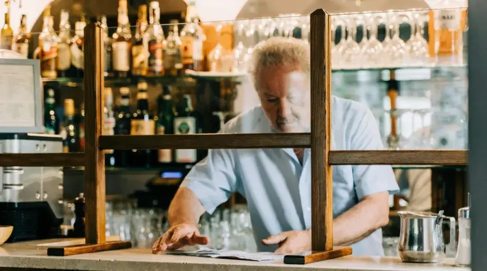 A small business owner standing behind the bar and protective sneeze guards prepares to open his bar.