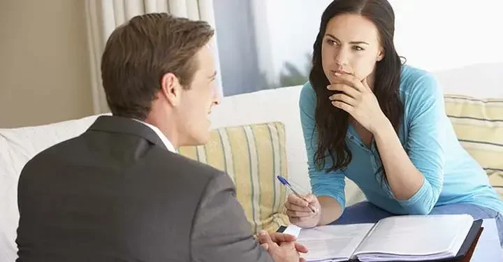 Woman in blue shirt with ballpoint pen hovering over documents looks at talking man in suit 