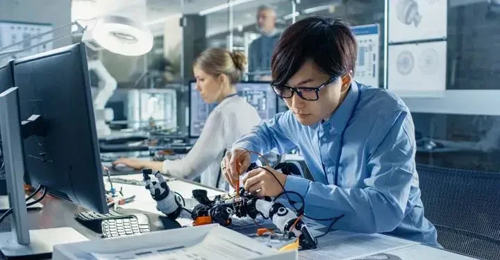 Man uses tools to adjust machine at desk in front of desktop computer