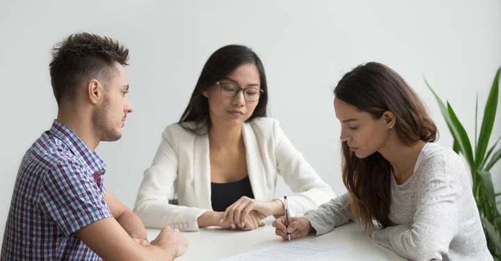 Two people sitting across from each other at a table signing documents while a separate woman sits at the head of the table, watching