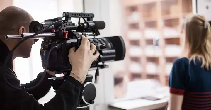 Bald male videographer holding camera pointed at blonde woman facing away