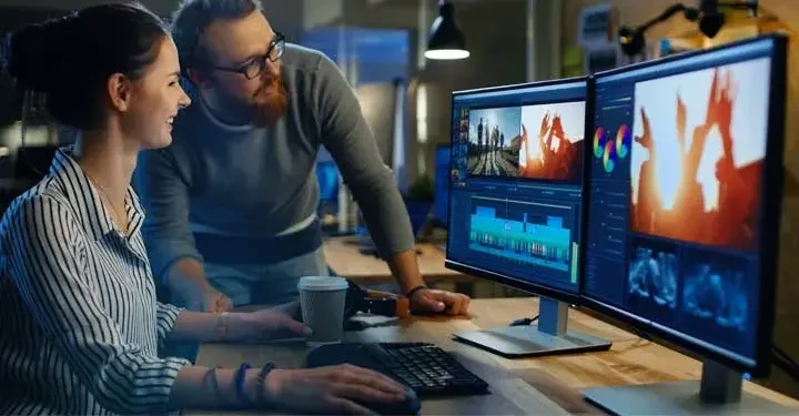 Man and woman editing videos on two desktop computers in low lit office