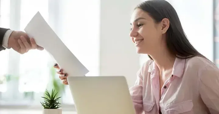 Woman working on laptop next to succulent is handed sheet of paper by person in suit