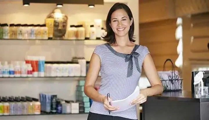 Smiling woman in front of bottled products holding papers and pens while leaning against granite counter