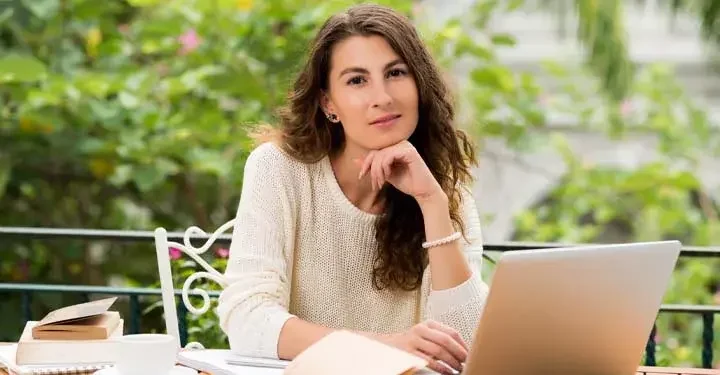 Woman resting chin on hand and typing on laptop sitting outside