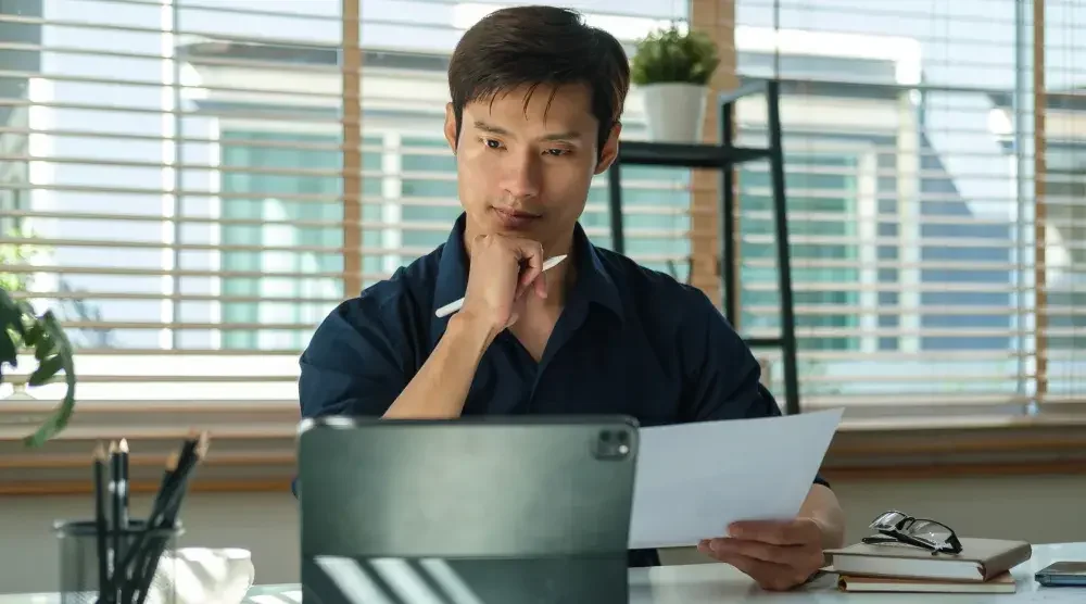 A man sits at his desk. He holds a document and reviews a business name search on his tablet.