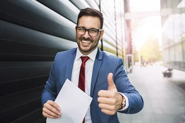 A smiling man in a suit carrying papers and giving the thumbs up.