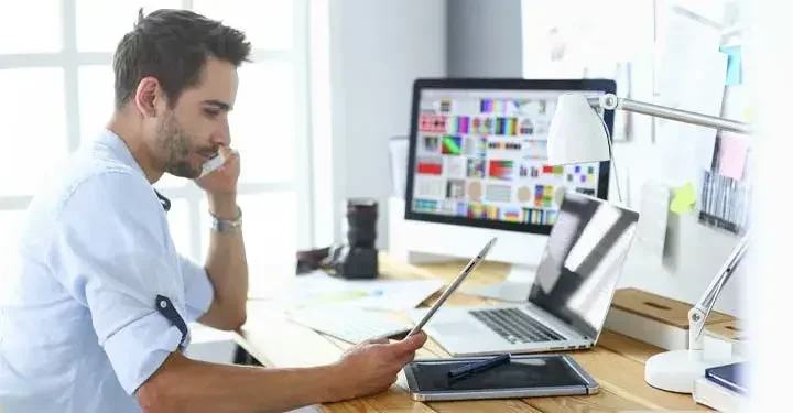 Man talking on cellphone holding tablet in an office with a laptop and a desktop on his table